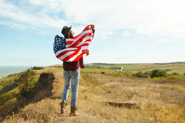 4th of July. Fourth of July. American with the national flag. American Flag. Independence Day. Patriotic holiday. The man is wearing a hat, a backpack, a shirt and jeans. Beautiful sunset light.