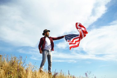 4 Temmuz. 4 Temmuz. Ulusal bayrağı olan Amerikalı. Amerikan bayrağı. Bağımsızlık günü. Vatanseverlik tatili. Adam şapka, sırt çantası, gömlek ve kot giyiyor. Güzel günbatımı ışık. 
