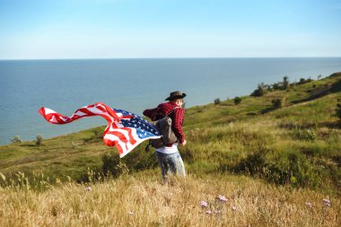 4 Temmuz. 4 Temmuz. Ulusal bayrağı olan Amerikalı. Amerikan bayrağı. Bağımsızlık günü. Vatanseverlik tatili. Adam şapka, sırt çantası, gömlek ve kot giyiyor. Güzel günbatımı ışık. 