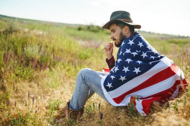 4 Temmuz. 4 Temmuz. Ulusal bayrağı olan Amerikalı. Amerikan bayrağı. Bağımsızlık günü. Vatanseverlik tatili. Adam şapka, sırt çantası, gömlek ve kot giyiyor. Güzel günbatımı ışık. 