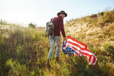 4 Temmuz. 4 Temmuz. Ulusal bayrağı olan Amerikalı. Amerikan bayrağı. Bağımsızlık günü. Vatanseverlik tatili. Adam şapka, sırt çantası, gömlek ve kot giyiyor. Güzel günbatımı ışık. 