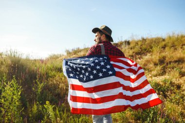 4 Temmuz. 4 Temmuz. Ulusal bayrağı olan Amerikalı. Amerikan bayrağı. Bağımsızlık günü. Vatanseverlik tatili. Adam şapka, sırt çantası, gömlek ve kot giyiyor. Güzel günbatımı ışık. 