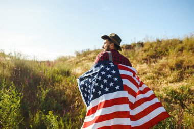 4 Temmuz. 4 Temmuz. Ulusal bayrağı olan Amerikalı. Amerikan bayrağı. Bağımsızlık günü. Vatanseverlik tatili. Adam şapka, sırt çantası, gömlek ve kot giyiyor. Güzel günbatımı ışık. 