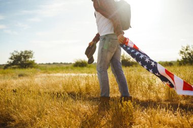 4 Temmuz. 4 Temmuz. Ulusal bayrağı olan Amerikalı. Amerikan bayrağı. Bağımsızlık günü. Vatanseverlik tatili. Adam şapka, sırt çantası, gömlek ve kot giyiyor. Güzel günbatımı ışık. 