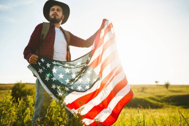 4 Temmuz. 4 Temmuz. Ulusal bayrağı olan Amerikalı. Amerikan bayrağı. Bağımsızlık günü. Vatanseverlik tatili. Adam şapka, sırt çantası, gömlek ve kot giyiyor. Güzel günbatımı ışık. 