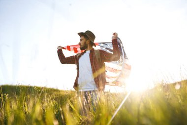 4 Temmuz. 4 Temmuz. Ulusal bayrağı olan Amerikalı. Amerikan bayrağı. Bağımsızlık günü. Vatanseverlik tatili. Adam şapka, sırt çantası, gömlek ve kot giyiyor. Güzel günbatımı ışık. 