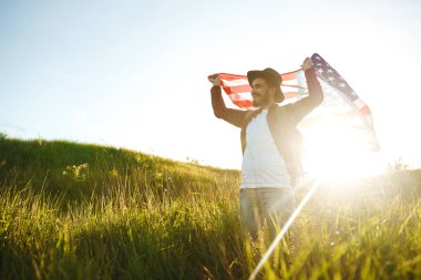 4 Temmuz. 4 Temmuz. Ulusal bayrağı olan Amerikalı. Amerikan bayrağı. Bağımsızlık günü. Vatanseverlik tatili. Adam şapka, sırt çantası, gömlek ve kot giyiyor. Güzel günbatımı ışık. 