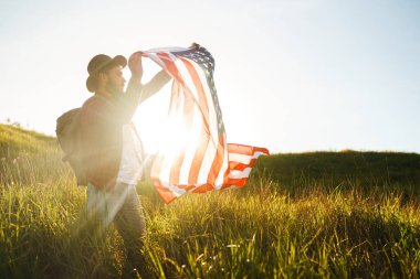 4 Temmuz. 4 Temmuz. Ulusal bayrağı olan Amerikalı. Amerikan bayrağı. Bağımsızlık günü. Vatanseverlik tatili. Adam şapka, sırt çantası, gömlek ve kot giyiyor. Güzel günbatımı ışık. 