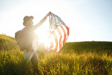 4 Temmuz. 4 Temmuz. Ulusal bayrağı olan Amerikalı. Amerikan bayrağı. Bağımsızlık günü. Vatanseverlik tatili. Adam şapka, sırt çantası, gömlek ve kot giyiyor. Güzel günbatımı ışık. 