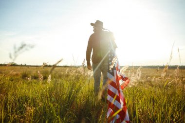 4 Temmuz. 4 Temmuz. Ulusal bayrağı olan Amerikalı. Amerikan bayrağı. Bağımsızlık günü. Vatanseverlik tatili. Adam şapka, sırt çantası, gömlek ve kot giyiyor. Güzel günbatımı ışık. 