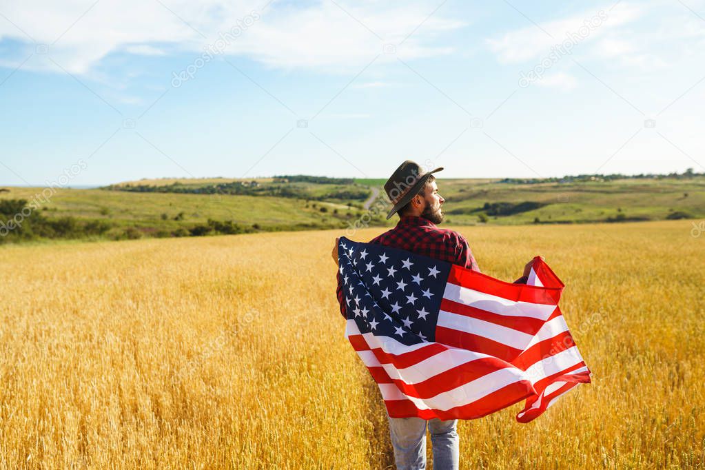 4th of July. Fourth of July. American with the national flag. American Flag. Independence Day. Patriotic holiday. The man is wearing a hat, a backpack, a shirt and jeans. Beautiful sunset light. 