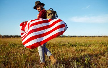 4 Temmuz. 4 Temmuz. Ulusal bayrağı olan Amerikalı. Amerikan bayrağı. Bağımsızlık günü. Vatanseverlik tatili. Adam şapka, sırt çantası, gömlek ve kot giyiyor. Güzel günbatımı ışık. 
