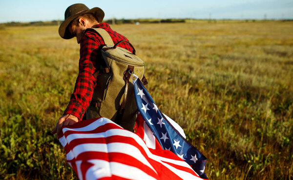 4th of July. Fourth of July. American with the national flag. American Flag. Independence Day. Patriotic holiday. The man is wearing a hat, a backpack, a shirt and jeans. Beautiful sunset light. 