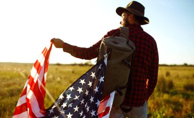 4 Temmuz. 4 Temmuz. Ulusal bayrağı olan Amerikalı. Amerikan bayrağı. Bağımsızlık günü. Vatanseverlik tatili. Adam şapka, sırt çantası, gömlek ve kot giyiyor. Güzel günbatımı ışık. 