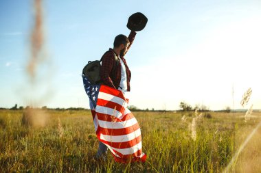 4 Temmuz. 4 Temmuz. Ulusal bayrağı olan Amerikalı. Amerikan bayrağı. Bağımsızlık günü. Vatanseverlik tatili. Adam şapka, sırt çantası, gömlek ve kot giyiyor. Güzel günbatımı ışık. 