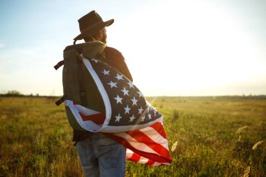 4 Temmuz. 4 Temmuz. Ulusal bayrağı olan Amerikalı. Amerikan bayrağı. Bağımsızlık günü. Vatanseverlik tatili. Adam şapka, sırt çantası, gömlek ve kot giyiyor. Güzel günbatımı ışık. 