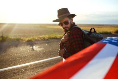 4 Temmuz. 4 Temmuz. Ulusal bayrağı olan Amerikalı. Amerikan bayrağı. Bağımsızlık günü. Vatanseverlik tatili. Adam şapka, sırt çantası, gömlek ve kot giyiyor. Güzel günbatımı ışık. 
