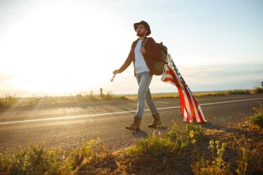 4 Temmuz. 4 Temmuz. Ulusal bayrağı olan Amerikalı. Amerikan bayrağı. Bağımsızlık günü. Vatanseverlik tatili. Adam şapka, sırt çantası, gömlek ve kot giyiyor. Güzel günbatımı ışık. 