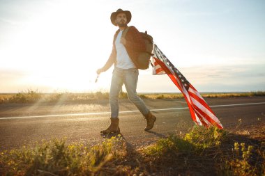 4 Temmuz. 4 Temmuz. Ulusal bayrağı olan Amerikalı. Amerikan bayrağı. Bağımsızlık günü. Vatanseverlik tatili. Adam şapka, sırt çantası, gömlek ve kot giyiyor. Güzel günbatımı ışık. 