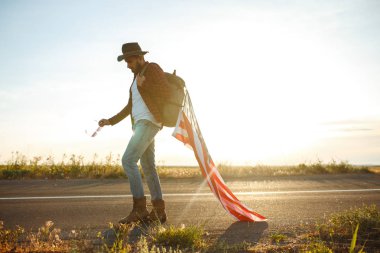 4 Temmuz. 4 Temmuz. Ulusal bayrağı olan Amerikalı. Amerikan bayrağı. Bağımsızlık günü. Vatanseverlik tatili. Adam şapka, sırt çantası, gömlek ve kot giyiyor. Güzel günbatımı ışık. 