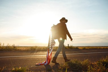 4 Temmuz. 4 Temmuz. Ulusal bayrağı olan Amerikalı. Amerikan bayrağı. Bağımsızlık günü. Vatanseverlik tatili. Adam şapka, sırt çantası, gömlek ve kot giyiyor. Güzel günbatımı ışık. 