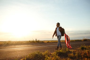4 Temmuz. 4 Temmuz. Ulusal bayrağı olan Amerikalı. Amerikan bayrağı. Bağımsızlık günü. Vatanseverlik tatili. Adam şapka, sırt çantası, gömlek ve kot giyiyor. Güzel günbatımı ışık. 