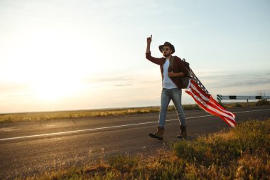 4 Temmuz. 4 Temmuz. Ulusal bayrağı olan Amerikalı. Amerikan bayrağı. Bağımsızlık günü. Vatanseverlik tatili. Adam şapka, sırt çantası, gömlek ve kot giyiyor. Güzel günbatımı ışık. 