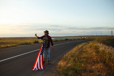4 Temmuz. 4 Temmuz. Ulusal bayrağı olan Amerikalı. Amerikan bayrağı. Bağımsızlık günü. Vatanseverlik tatili. Adam şapka, sırt çantası, gömlek ve kot giyiyor. Güzel günbatımı ışık. 