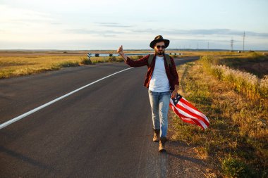 4 Temmuz. 4 Temmuz. Ulusal bayrağı olan Amerikalı. Amerikan bayrağı. Bağımsızlık günü. Vatanseverlik tatili. Adam şapka, sırt çantası, gömlek ve kot giyiyor. Güzel günbatımı ışık. 