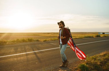 4 Temmuz. 4 Temmuz. Ulusal bayrağı olan Amerikalı. Amerikan bayrağı. Bağımsızlık günü. Vatanseverlik tatili. Adam şapka, sırt çantası, gömlek ve kot giyiyor. Güzel günbatımı ışık. 