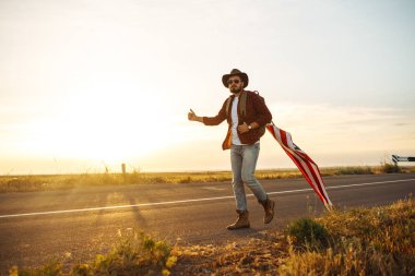 4 Temmuz. 4 Temmuz. Ulusal bayrağı olan Amerikalı. Amerikan bayrağı. Bağımsızlık günü. Vatanseverlik tatili. Adam şapka, sırt çantası, gömlek ve kot giyiyor. Güzel günbatımı ışık. 