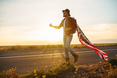 4 Temmuz. 4 Temmuz. Ulusal bayrağı olan Amerikalı. Amerikan bayrağı. Bağımsızlık günü. Vatanseverlik tatili. Adam şapka, sırt çantası, gömlek ve kot giyiyor. Güzel günbatımı ışık. 