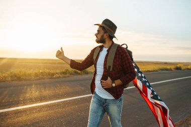 4 Temmuz. 4 Temmuz. Ulusal bayrağı olan Amerikalı. Amerikan bayrağı. Bağımsızlık günü. Vatanseverlik tatili. Adam şapka, sırt çantası, gömlek ve kot giyiyor. Güzel günbatımı ışık. 