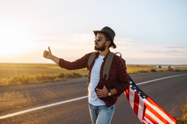 4 Temmuz. 4 Temmuz. Ulusal bayrağı olan Amerikalı. Amerikan bayrağı. Bağımsızlık günü. Vatanseverlik tatili. Adam şapka, sırt çantası, gömlek ve kot giyiyor. Güzel günbatımı ışık. 