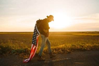4 Temmuz. 4 Temmuz. Ulusal bayrağı olan Amerikalı. Amerikan bayrağı. Bağımsızlık günü. Vatanseverlik tatili. Adam şapka, sırt çantası, gömlek ve kot giyiyor. Güzel günbatımı ışık. 
