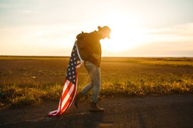 4 Temmuz. 4 Temmuz. Ulusal bayrağı olan Amerikalı. Amerikan bayrağı. Bağımsızlık günü. Vatanseverlik tatili. Adam şapka, sırt çantası, gömlek ve kot giyiyor. Güzel günbatımı ışık. 