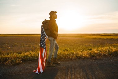 4 Temmuz. 4 Temmuz. Ulusal bayrağı olan Amerikalı. Amerikan bayrağı. Bağımsızlık günü. Vatanseverlik tatili. Adam şapka, sırt çantası, gömlek ve kot giyiyor. Güzel günbatımı ışık. 