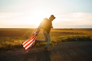 4 Temmuz. 4 Temmuz. Ulusal bayrağı olan Amerikalı. Amerikan bayrağı. Bağımsızlık günü. Vatanseverlik tatili. Adam şapka, sırt çantası, gömlek ve kot giyiyor. Güzel günbatımı ışık. 