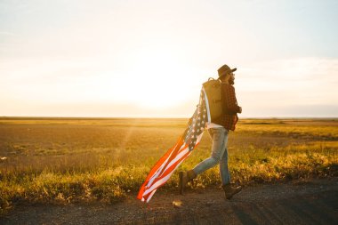 4 Temmuz. 4 Temmuz. Ulusal bayrağı olan Amerikalı. Amerikan bayrağı. Bağımsızlık günü. Vatanseverlik tatili. Adam şapka, sırt çantası, gömlek ve kot giyiyor. Güzel günbatımı ışık. 