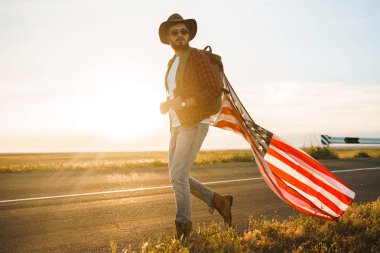 4 Temmuz. 4 Temmuz. Ulusal bayrağı olan Amerikalı. Amerikan bayrağı. Bağımsızlık günü. Vatanseverlik tatili. Adam şapka, sırt çantası, gömlek ve kot giyiyor. Güzel günbatımı ışık. 