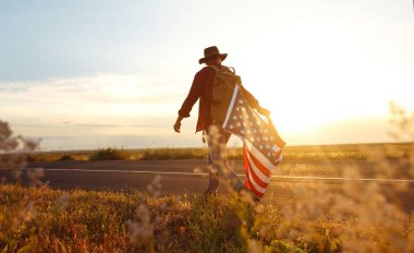 4 Temmuz. 4 Temmuz. Ulusal bayrağı olan Amerikalı. Amerikan bayrağı. Bağımsızlık günü. Vatanseverlik tatili. Adam şapka, sırt çantası, gömlek ve kot giyiyor. Güzel günbatımı ışık. 