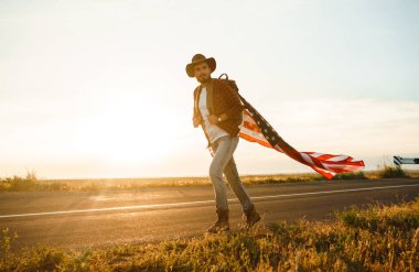 4 Temmuz. 4 Temmuz. Ulusal bayrağı olan Amerikalı. Amerikan bayrağı. Bağımsızlık günü. Vatanseverlik tatili. Adam şapka, sırt çantası, gömlek ve kot giyiyor. Güzel günbatımı ışık. 