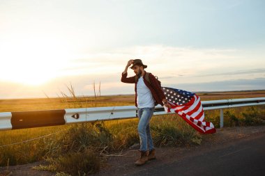 4 Temmuz. 4 Temmuz. Ulusal bayrağı olan Amerikalı. Amerikan bayrağı. Bağımsızlık günü. Vatanseverlik tatili. Adam şapka, sırt çantası, gömlek ve kot giyiyor. Güzel günbatımı ışık. 