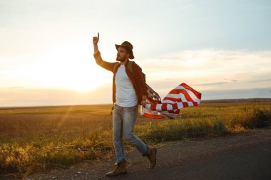 4 Temmuz. 4 Temmuz. Ulusal bayrağı olan Amerikalı. Amerikan bayrağı. Bağımsızlık günü. Vatanseverlik tatili. Adam şapka, sırt çantası, gömlek ve kot giyiyor. Güzel günbatımı ışık. 