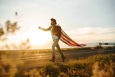4 Temmuz. 4 Temmuz. Ulusal bayrağı olan Amerikalı. Amerikan bayrağı. Bağımsızlık günü. Vatanseverlik tatili. Adam şapka, sırt çantası, gömlek ve kot giyiyor. Güzel günbatımı ışık. 