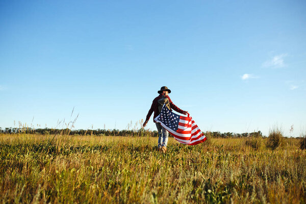 4 июля. Четвертое июля. Американец с национальным флагом. Американский флаг. День независимости. Патриотический праздник. На человеке шляпа, рюкзак, рубашка и джинсы. Закат света
. 