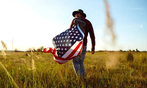 4th of July. Fourth of July. American with the national flag. American Flag. Independence Day. Patriotic holiday. The man is wearing a hat, a backpack, a shirt and jeans. Beautiful sunset light.