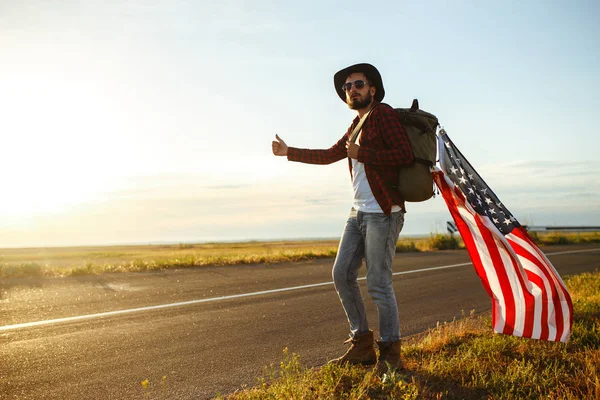 4 Temmuz. 4 Temmuz. Ulusal bayrağı olan Amerikalı. Amerikan bayrağı. Bağımsızlık günü. Vatanseverlik tatili. Adam şapka, sırt çantası, gömlek ve kot giyiyor. Güzel günbatımı ışık. 
