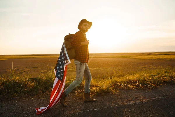 4th of July. Fourth of July. American with the national flag. American Flag. Independence Day. Patriotic holiday. The man is wearing a hat, a backpack, a shirt and jeans. Beautiful sunset light.