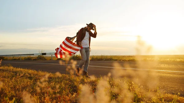 4 Temmuz. 4 Temmuz. Ulusal bayrağı olan Amerikalı. Amerikan bayrağı. Bağımsızlık günü. Vatanseverlik tatili. Adam şapka, sırt çantası, gömlek ve kot giyiyor. Güzel günbatımı ışık. 
